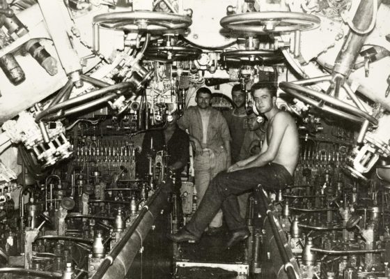Cinq jeunes hommes, membre de l'équipage du sous-marin Dauphin, posent dans l'espace des moteurs diesel du sous-marin. Le premier est torse nu.