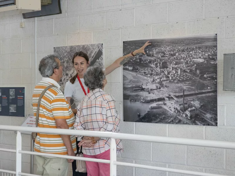 Photographe : Chloé Demora. Saint-Nazaire Agglomération Tourisme. Une médiatrice de l'Écomusée présente l'exposition photographique temporaire "Saint-Nazaire, vue d'en haut" à des visiteurs.