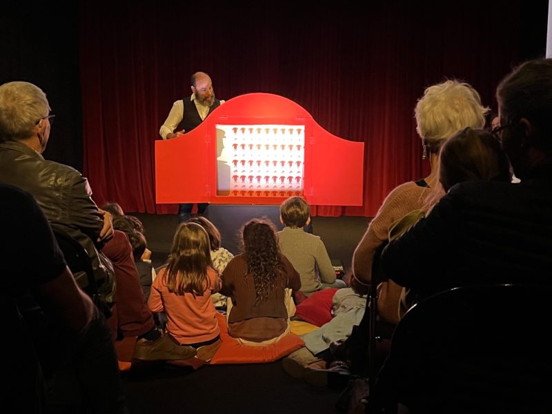 Photographie d'un spectacle de kamishibaï avec des enfants de dos et un comédien animant un petit théâtre.