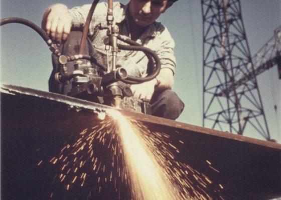 Photographie couleur d'un ouvrier accroupi sur une tôle en train de la découper et faisant de nombreuses étincelles.