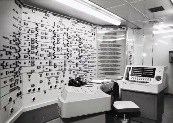 Photographe : Philippe Baudry. Collection Saint-Nazaire Agglomération Tourisme - Écomusée. Fonds Chantiers de l’Atlantique. Photographie noir et blanc du central sécurité du paquebot France (1962) avec un mur recouvert de voyants lumineux et un autre représentant toutes les ponts du navire.