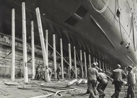 Photographe : inconnu. Collection Saint-Nazaire Agglomération Tourisme - Écomusée. Fonds Chantiers de l’Atlantique. Photographie en noir et blanc d'un groupe d'ouvriers tirant sur une corde pour faire tomber un pieu de bois sur lequel repose le paquebot France.