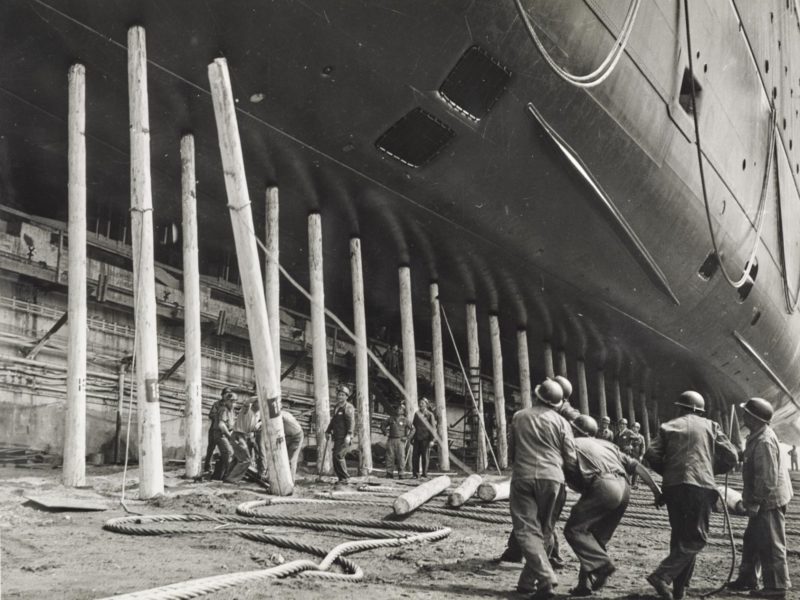 Photographie en noir et blanc d'un groupe d'ouvriers tirant sur une corde pour faire tomber un pieu de bois sur lequel repose le paquebot France.