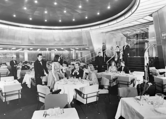 Photographe : inconnu. Collection Saint-Nazaire Agglomération Tourisme - Écomusée. Fonds Chantiers de l’Atlantique. Photographie noir et blanc représentant des passagers dans la salle à manger 1re classe, dite Chambord, du paquebot France (1962).