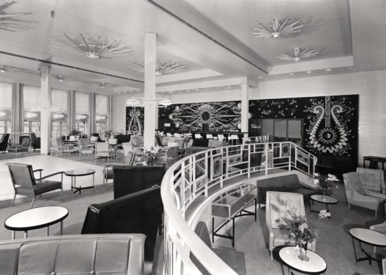 Photographie noir et blanc du fumoir 1re classe du paquebot France (1962), conçu par le décorateur André Arbus avec des canapés, des fauteuils, des tables basses et le mur du fond recouvert par la tapisserie Soleil d'Orphée de Picart-Ledoux.