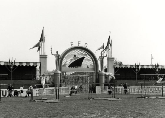 Deux jeunes femmes sont en train de faire des figures de gymnastique sur des barres parallèles dans le stade du Plessis devant le portail d'entrée représentant le paquebot France.