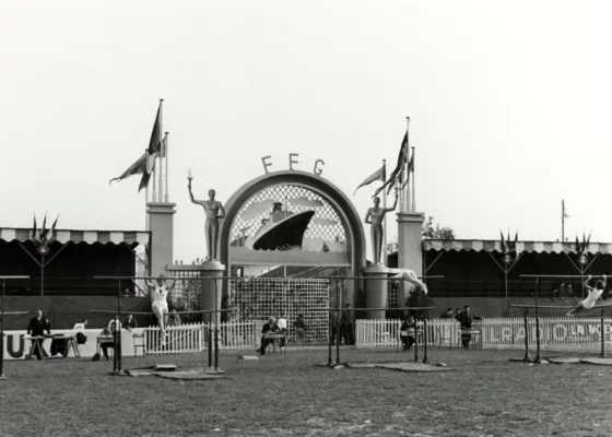 Deux jeunes femmes sont en train de faire des figures de gymnastique sur des barres parallèles dans le stade du Plessis devant le portail d'entrée représentant le paquebot France.
