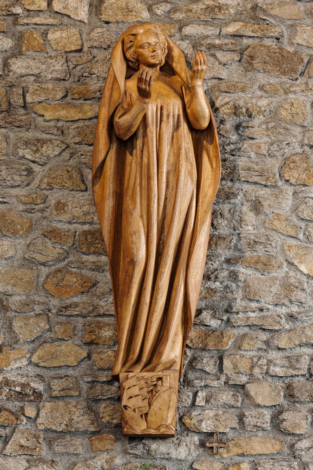 Sculpture en bois représentant une femme debout levant les mains au ciel, vêtue d'une longue robe et d'une cape, à ses pieds un chapiteau sculpté représentant un navire à voile et un paquebot.