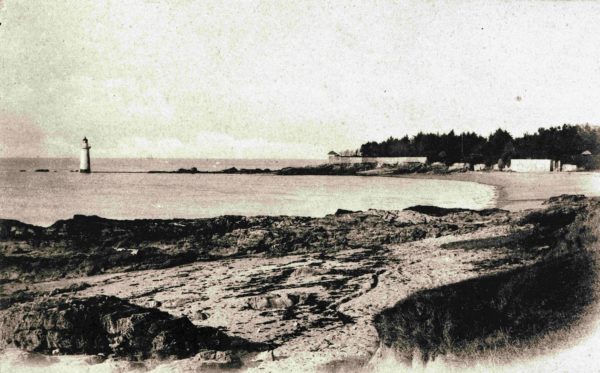 Carte postale de la plage de Villès-Martin. Au premier plan, la plage et ses rochers. Le phare de Villès-Martin est installé au bout d'une digue rocheuse. Au fond, une forêt et des habitations bordent la plage. À l'horizon, un voilier est perceptible.
