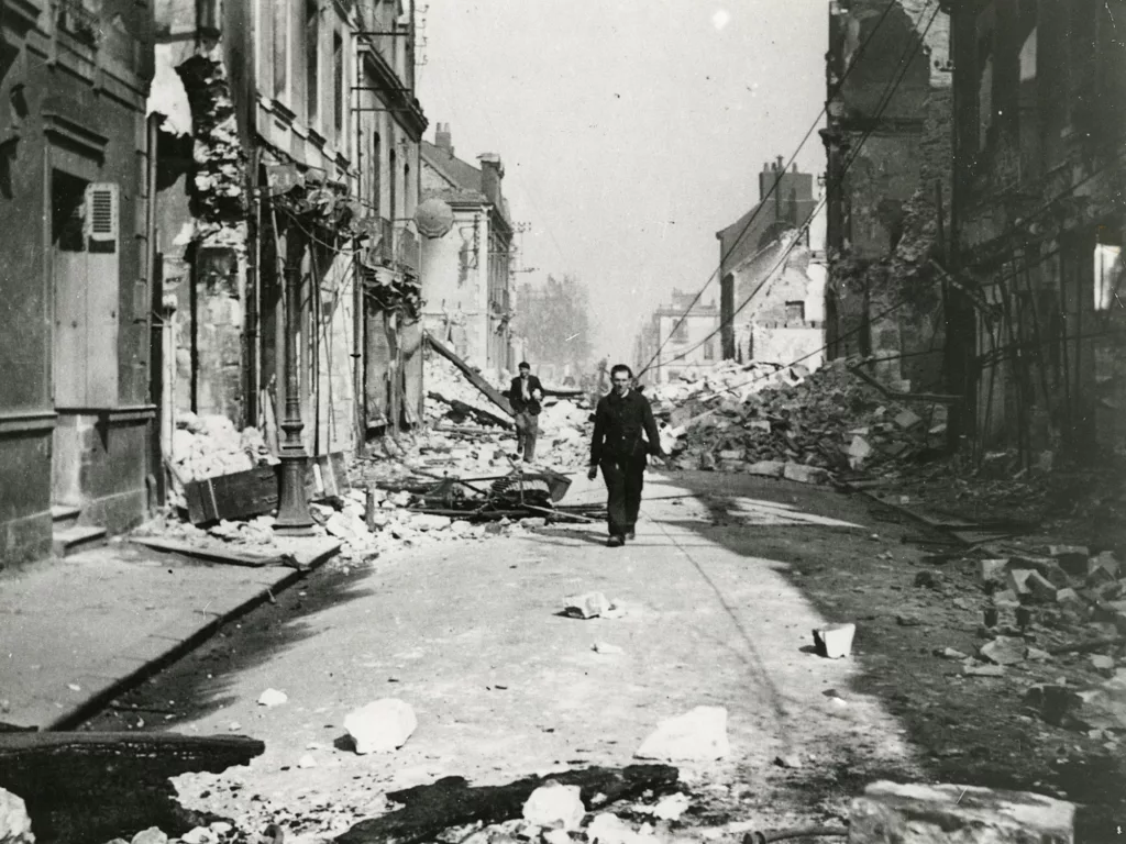 Photographie noir et blanc d'un homme qui marche face à l'objectif dans une rue en ruine de Saint-Nazaire pendant la Seconde Guerre mondiale.