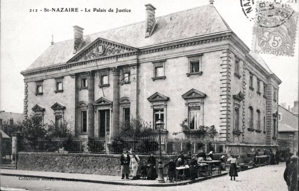 Carte postale noir et blanc du palais de justice de Saint-Nazaire, un jour de marché, vers 1900. Hommes, femmes et enfants regardent l'objectif depuis le trottoir devant le bâtiment.