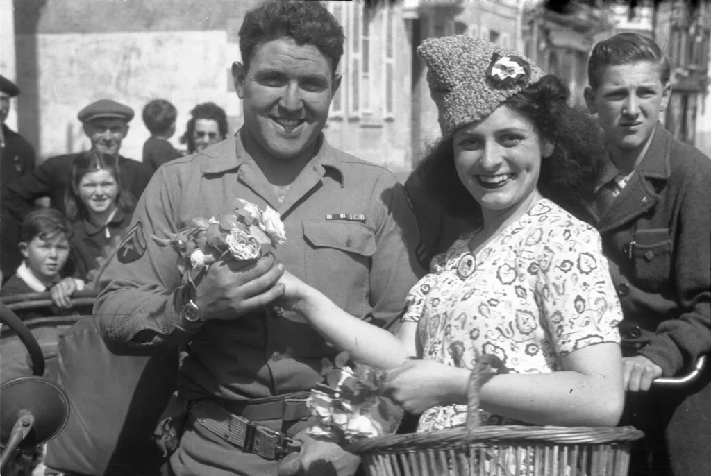 Photographie de la libération de la Poche Saint-Nazaire montrant un soldat américain souriant recevant des fleurs d'une jeune femme française en robe dans une rue bondée.