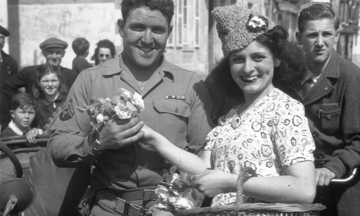 Photographie de la libération de la Poche Saint-Nazaire montrant un soldat américain souriant recevant des fleurs d'une jeune femme française en robe dans une rue bondée.