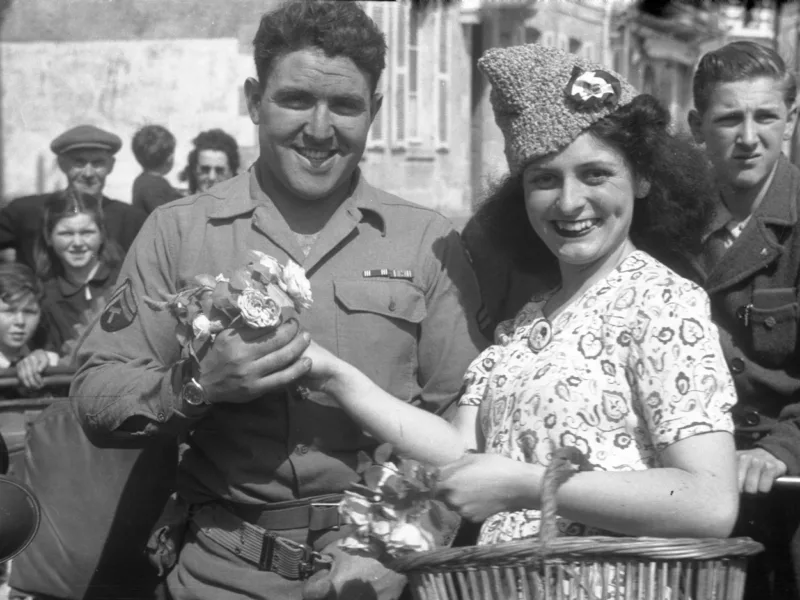 Photographie de la libération de la Poche Saint-Nazaire montrant un soldat américain souriant recevant des fleurs d'une jeune femme française en robe dans une rue bondée.