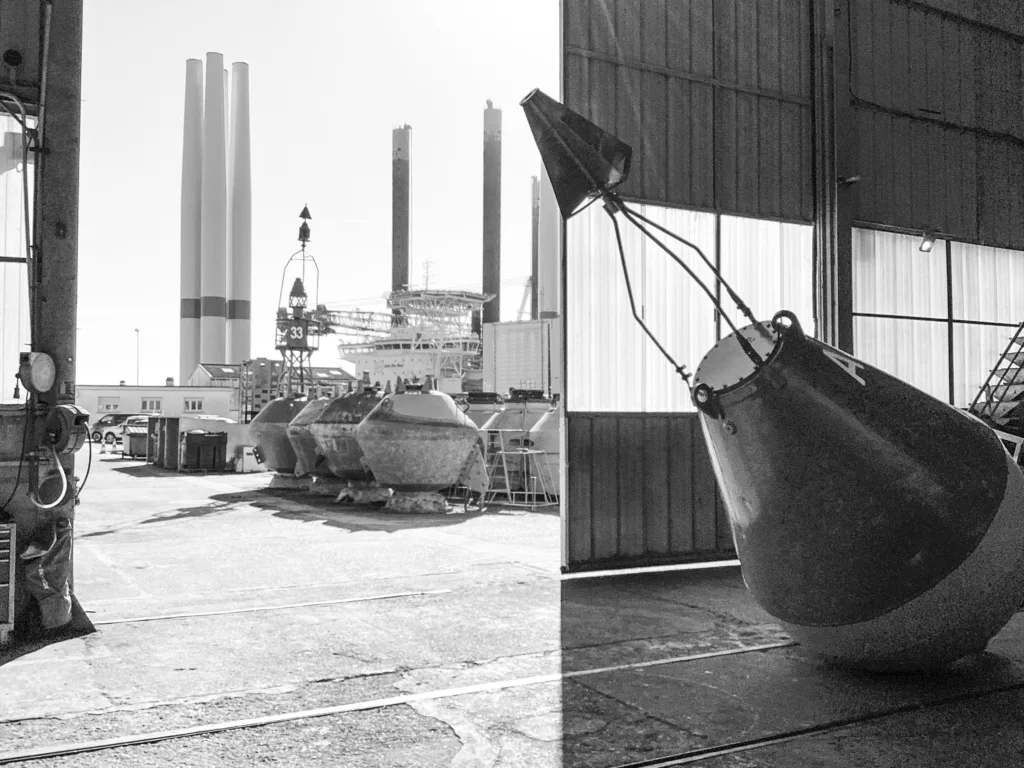 Photographes : Élèves de 3eF – Collège Anita Conti © Centre photographique Hasy Vue en noir et blanc de l'atelier des Phares et Balises à Saint-Nazaire. Une grande bouée métallique est stockée à l'intérieur d'un hangar ouvert, tandis que d'autres bouées et un navire de maintenance sont visibles sur le quai ensoleillé en arrière-plan.