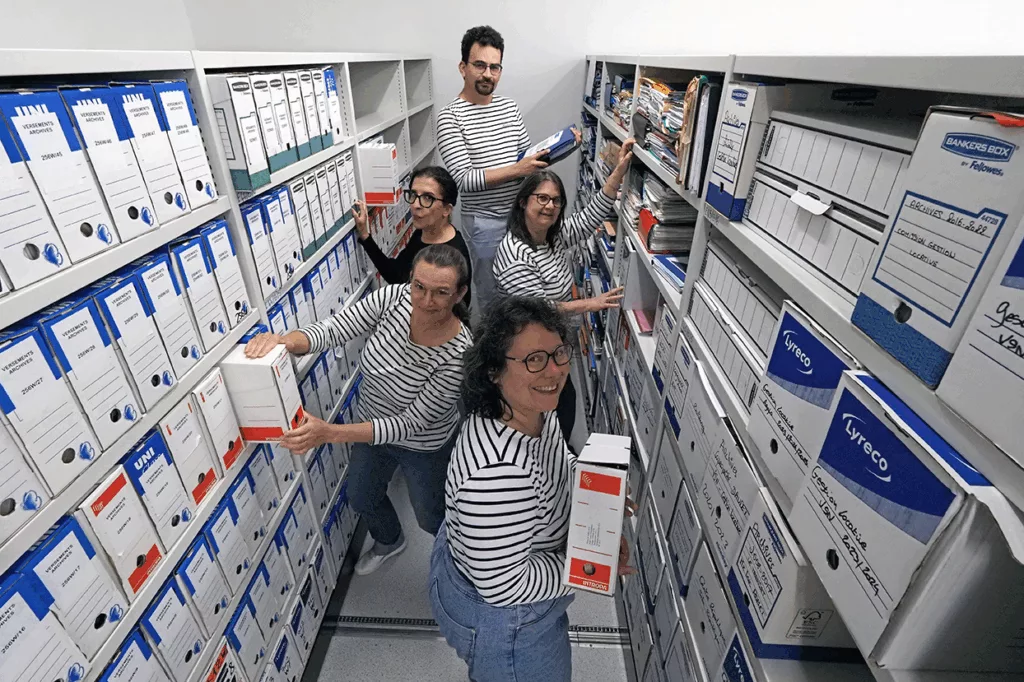 Vue en plongée de cinq membres de l'équipe souriants au milieu d'une allée de rayonnages remplis de boîtes d'archives. Plusieurs professionnels portent des hauts à rayures de style marinière.