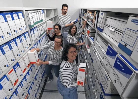 Vue en plongée de cinq membres de l'équipe souriants au milieu d'une allée de rayonnages remplis de boîtes d'archives. Plusieurs professionnels portent des hauts à rayures de style marinière.