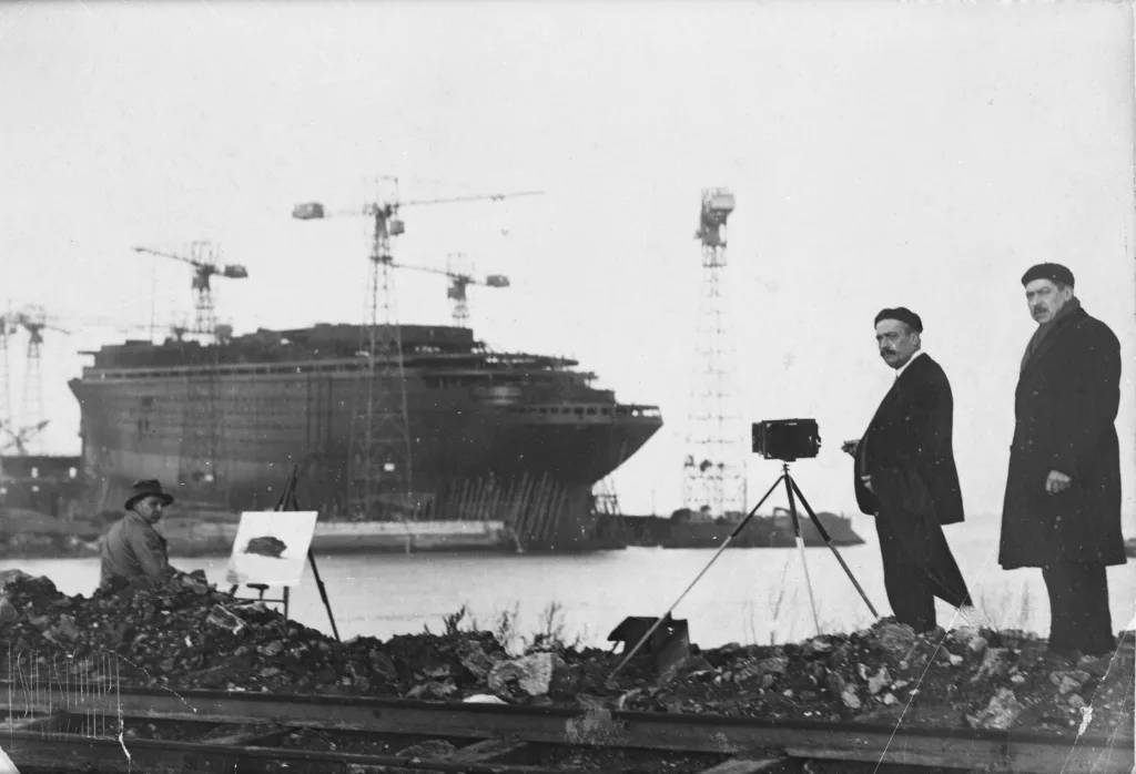 Photographe : inconnu. Collection particulière : Auguste Letouzo. Cliché Saint-Nazaire Agglomération Tourisme - Écomusée. Photographie en noir et blanc de la construction du paquebot Normandie à Saint-Nazaire. Un peintre et un photographe immortalisent le navire sur sa cale de lancement