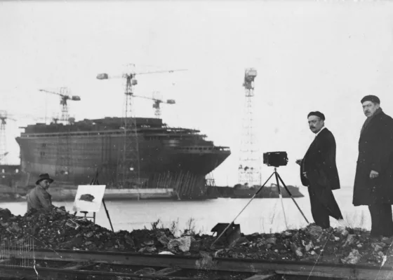 Photographie en noir et blanc de la construction du paquebot Normandie à Saint-Nazaire. Un peintre et un photographe immortalisent le navire sur sa cale de lancement