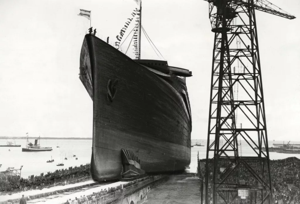 Photographe : inconnu. Collection Saint-Nazaire Agglomération Tourisme - Écomusée. Fonds François Blancho. Photographie en noir et blanc du lancement du paquebot Normandie aux chantiers navals de Saint-Nazaire, vue depuis la cale de construction avec une grue géante à droite. La foule est assemblée pour assister à cet événement d'ampleur.