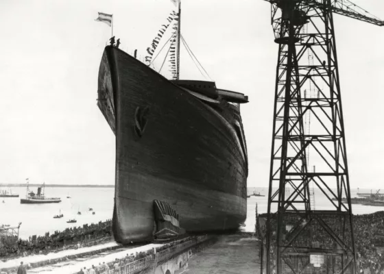 Photographie en noir et blanc du lancement du paquebot Normandie aux chantiers navals de Saint-Nazaire, vue depuis la cale de construction avec une grue géante à droite. La foule est assemblée pour assister à cet événement d'ampleur.